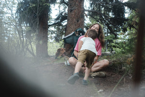 Little boy running toward his smiling mother for a hug during a hiking break in the forest, with their dog relaxing beside them.