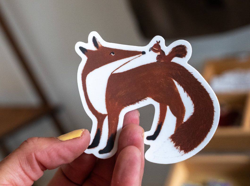 Hand holding a sticker of a red fox with a squirrel riding on its back, illustrated in earthy tones.