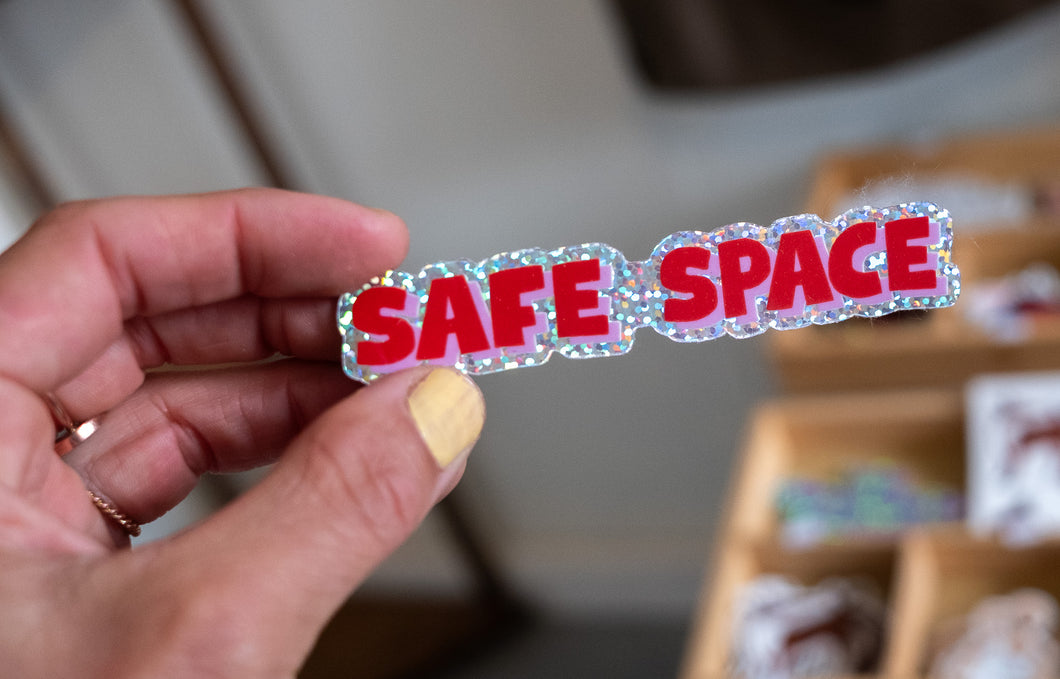 Hand holding a glittery sticker with bold red letters reading “Safe Space.”