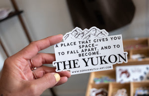 Hand holding a black and white sticker with a mountain illustration and the quote: “A place that gives you space—to fall apart, and to become. THE YUKON.”