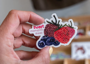 Hand holding a colorful fruit sticker that reads “sweet moments ahead,” featuring strawberries and blueberries.