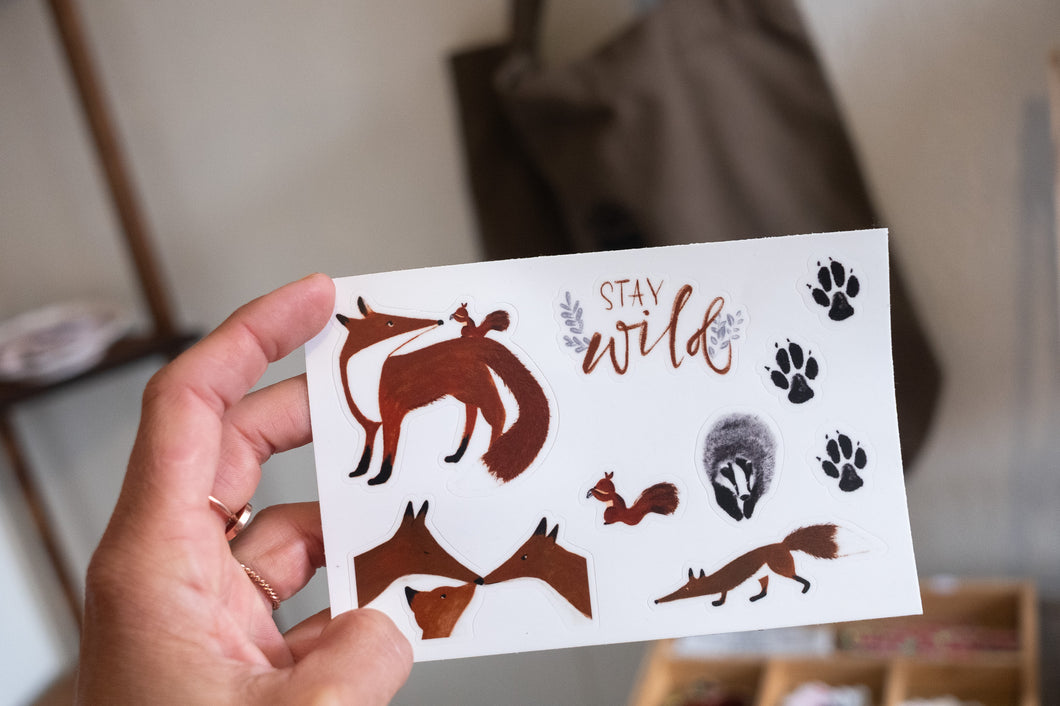 Hand holding a fox-themed sticker sheet with woodland illustrations, paw prints, and the phrase “stay wild.”