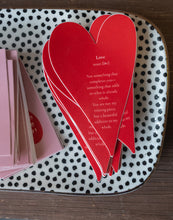 Stack of bright red heart-shaped stickers with the word “Love” and a poetic definition, placed on a spotted tray.