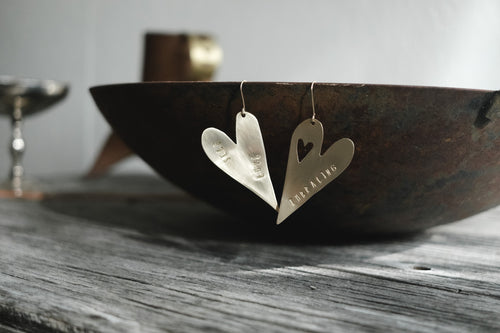 Brass heart earrings stamped with “Self Love” and “Embracing” resting against a rustic metal bowl.